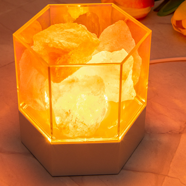 Modern Himalayan salt lamp lit close up, rough pink salt chunks glowing inside the clear hexagonal case on its white base, USB cord visible