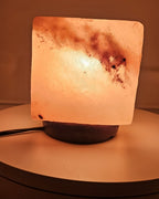 Himalayan salt cube lamp lit from the front, on a turned base with the power cord visible