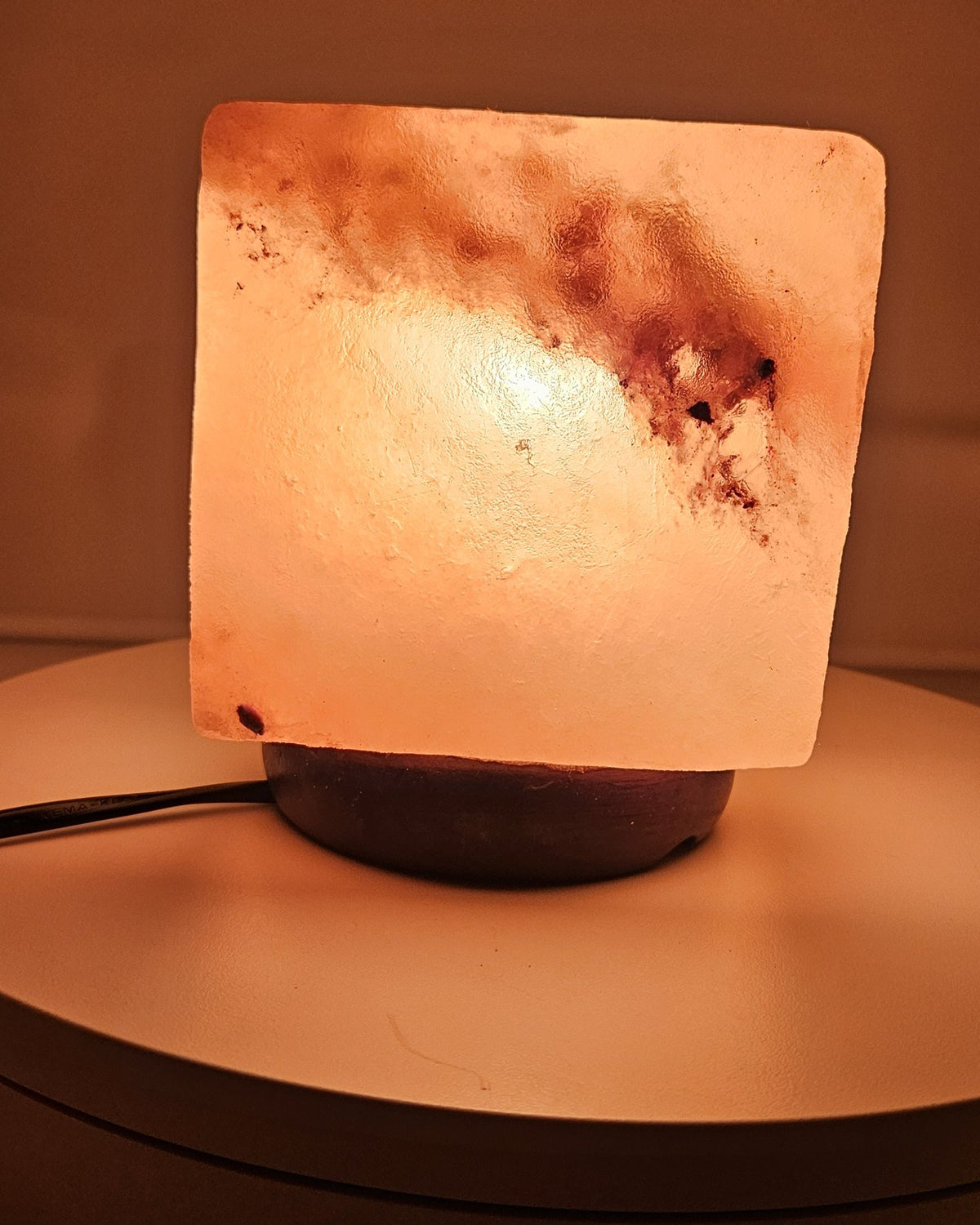 Himalayan salt cube lamp lit from the front, on a turned base with the power cord visible