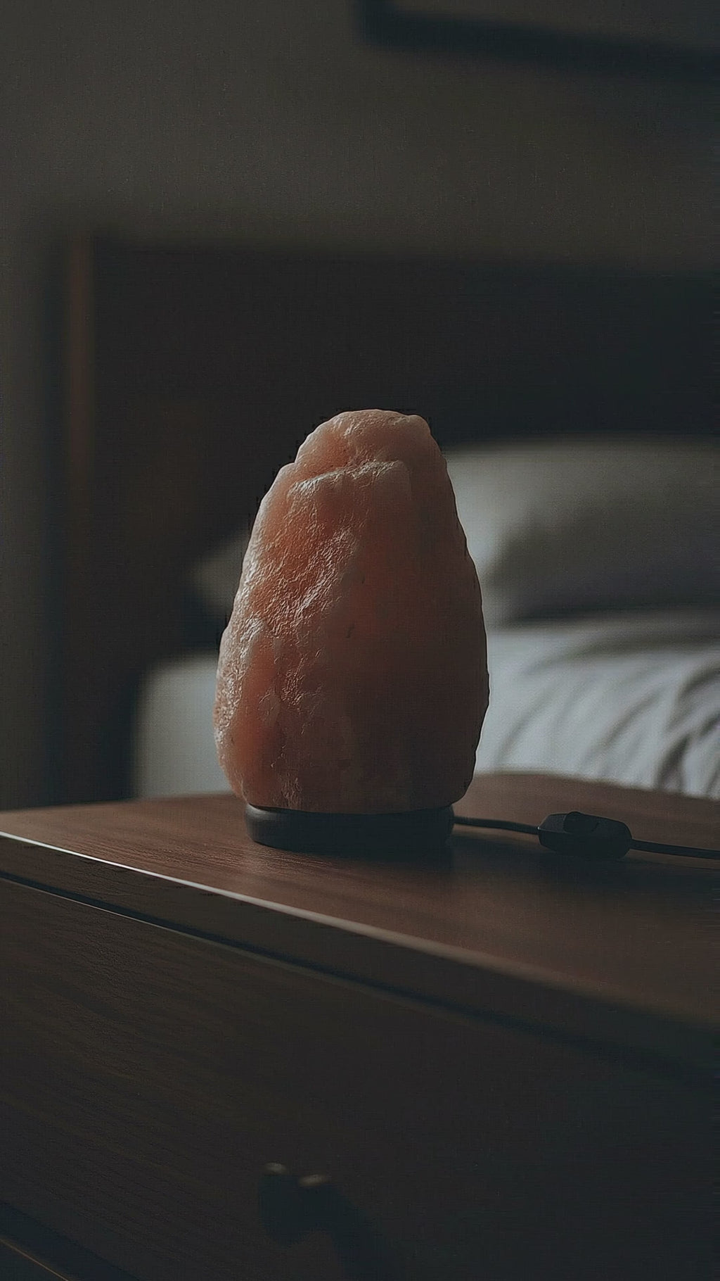 Natural Himalayan pink salt lamp warming on in a dark bedroom until its crystal fills with a soft amber glow, cord and inline switch visible