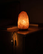 Himalayan salt lamp dimmed to a low ember glow in a dark bedroom, the inline dimmer on its black cord catching the light