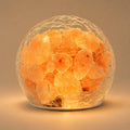 Battery powered mini Himalayan salt lamp switched on, rough pink salt pieces glowing warm amber inside a clear crackle-finish glass globe on a small white base