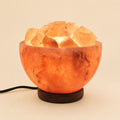 Himalayan salt bowl lamp filled with lit salt chunks on a dark wooden base, power cord visible
