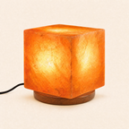 Cube Himalayan salt lamp lit on a round natural wooden base, power cord visible